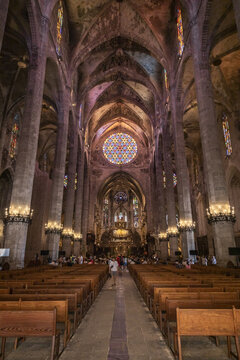View of the interior of the Palma Cathedral with its towering stone columns, intricate vaulted ceilings, and a vibrant stained glass rose window above the nave in Palma, Balearic Islands, Spain.