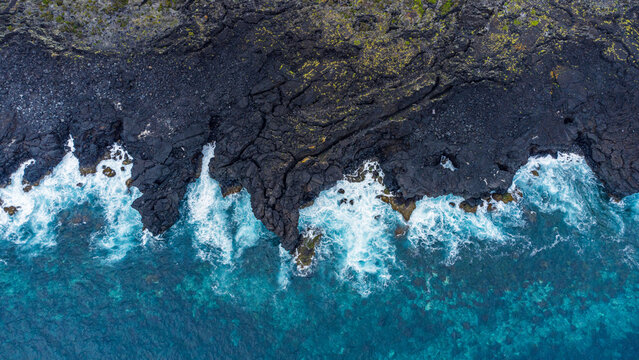 Aerial view of rugged black volcanic lava rock coastline meeting turquoise ocean waves, Azores, Portugal.