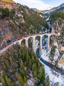 Aerial view of the Landwasser Viaduct with the red Glacier express crossing the stone arch bridge over a river in a snowy mountain landscape in Grisons, Switzerland.