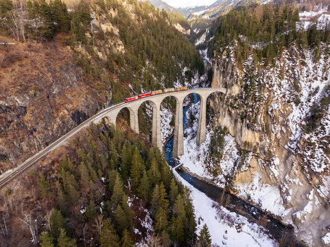 Aerial view of the Landwasser Viaduct with a train crossing the stone arch bridge over a snowy gorge in Grisons, Switzerland.