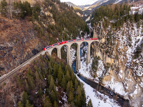 Aerial view of the Landwasser Viaduct with the red Glacier express crossing the stone arch bridge over a snowy valley with a river and pine trees in Grisons, Switzerland.