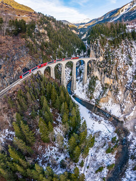 Aerial view of the Landwasser Viaduct, its stone arches carrying the red Glacier Express, framed by snow-covered mountains, pine forests, and a winding river in Grisons, Switzerland.