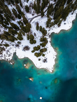 Aerial view of a turquoise mountain lake shore covered in fresh snow with dense evergreen pine trees and a winding road in Grisons, Switzerland.