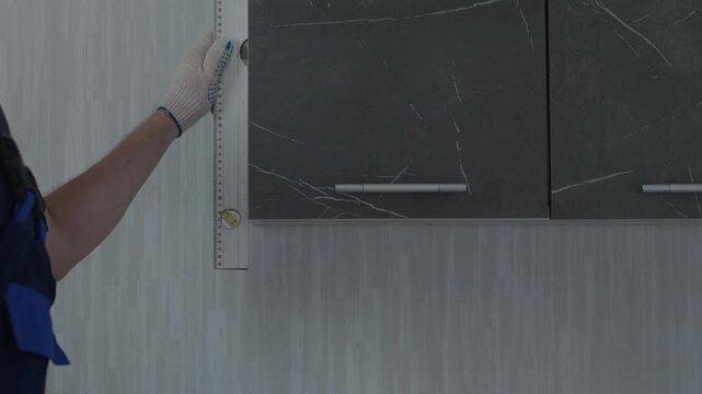 A professional handyman uses a spirit level to check the vertical alignment of a newly installed kitchen cabinet. The worker measures the precision of the modern dark wooden furniture 