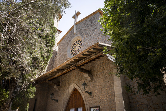 View of the stone facade of the church of Sant Ramon de Penyafort with its rose window and wooden entrance framed by lush trees in Port de Soller, Balearic Islands, Spain.