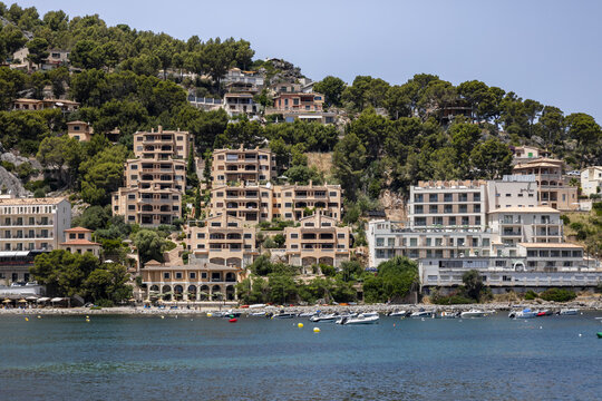 View of the coastal town of Port de Soller with its hillside hotels, lush pine trees, and small boats anchored in the bay, Port de Soller, Balearic Islands, Spain.