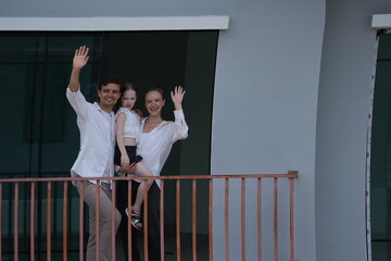 Fototapeta premium Caucasian family of three embracing wave happily from their balcony, the girl riding piggyback on the balcony of their new house overlooking the residential estate, authentic everyday scene
