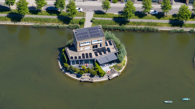 Aerial view of a modern house with solar panels on a small circular island in a canal, featuring a deck, garden, and people, in Heerhugowaard, North Holland, Netherlands.