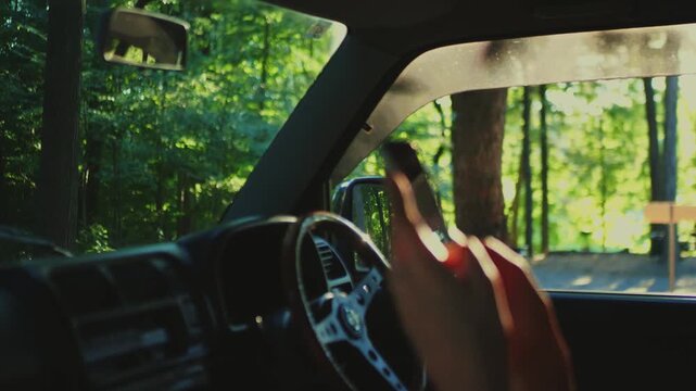Close-up of a hand holding a smartphone inside a car parked near a sunlit forest road. Represents travel,  communication, and road trip lifestyle.