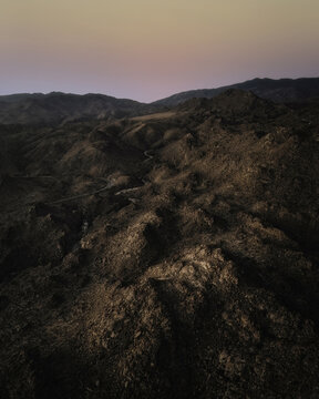 Aerial view of the rugged, rocky desert landscape of Southern California at sunset, featuring a winding road through the hills under a soft pink sky, California, United States.