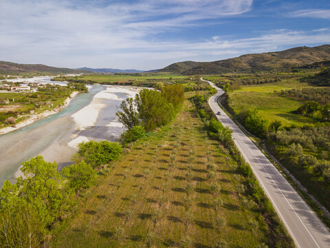 Aerial view of the Drashovica landscape featuring a winding river, a paved road, and rows of olive trees in the fields under a clear blue sky in Drashovica, Vlores, Albania.