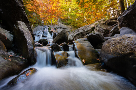View of the Lagoni waterfall cascading over large mossy rocks surrounded by vibrant autumn foliage in the forest of Corniglio, Emilia-Romagna, Italy.