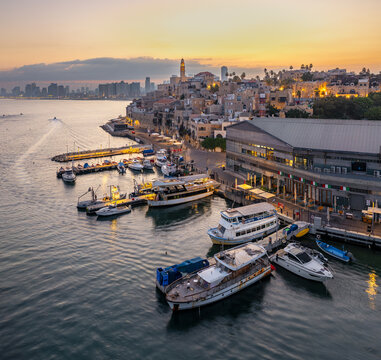 Aerial view of Jaffa Port with boats docked at the pier, historic stone buildings, and the Tel Aviv skyline under a warm sunset sky in Tel Aviv-Jaffa, Tel Aviv District, Israel.