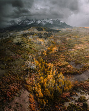 Aerial view of the Million Dollar Highway winding through yellow aspen trees and autumn foliage, set against snow-capped mountains under a stormy sky in Ouray, Colorado, United States.