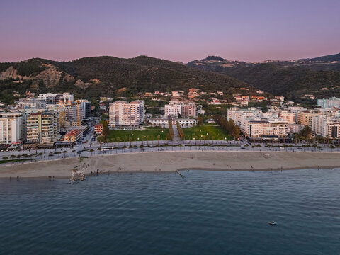 Aerial view of the coastal city of Vlora at sunset, featuring a sandy beach, promenade, modern apartment buildings, and rolling hills in the background, Vlora, Vlore County, Albania.