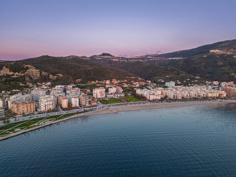 Aerial view of the coastal city with modern buildings, sandy beach, and green parks nestled against rolling mountains under a twilight sky in Vlora, Vlore County, Albania.
