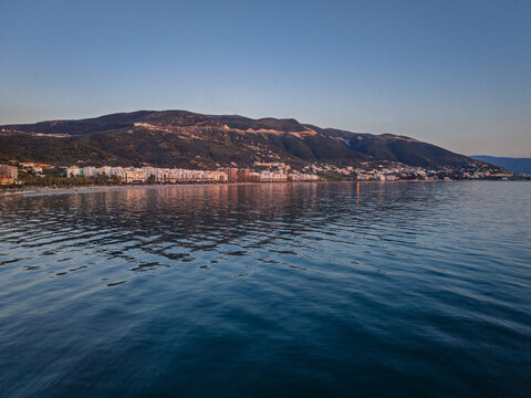 Aerial view of the Vlora coastline at sunset with calm sea, city buildings along the shore, and mountains in the background in Vlora, Vlore County, Albania.