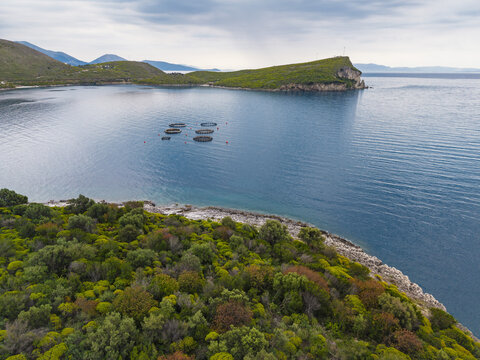 Aerial view of Porto Palermo featuring lush green coastal vegetation, rocky shoreline, and fish farming cages floating in the calm blue sea, Porto Palermo, Qarku i Vlores, Albania.