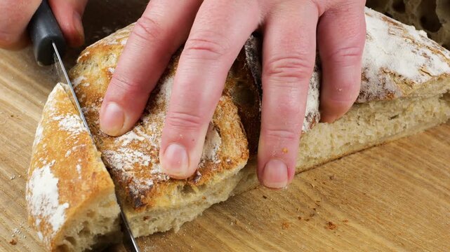 A serrated knife slices freshly baked homemade sourdough wheat bread on a wooden board. Close-up captures the crusty artisan loaf being cut into slices, showing its traditional texture. Vertical video