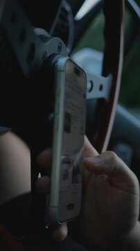 Close-up of tattooed hands scrolling or using a smartphone inside a car, with the steering wheel and dashboard. Screen is blurred. Mobile communication, digital technology, and online shopping. 4K.