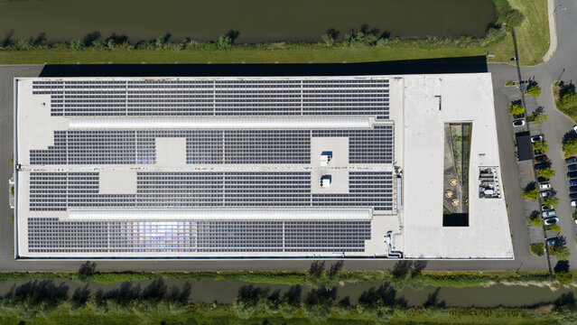 Aerial view of a large industrial building with extensive solar panel arrays on its flat roof, situated next to a canal and parking area in Houten, Utrecht, Netherlands.