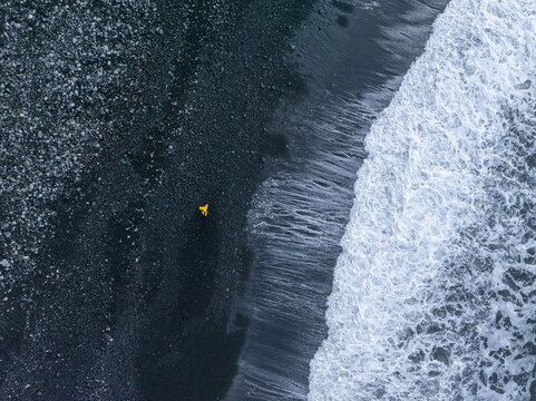 Aerial view of a person in a yellow jacket walking on the dark volcanic pebble beach of Faja de Lopo Vaz in Lajes das Flores, Azores, Portugal.