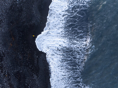 Aerial view of Faja de Lopo Vaz with a person in a yellow jacket walking along the black sand beach as waves crash against the shore in Lajes das Flores, Azores, Portugal.