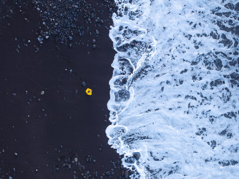 Aerial view of Faja de Lopo Vaz with a person in a yellow jacket standing on the black sand beach as ocean waves crash against the shore in Lajes das Flores, Azores, Portugal.