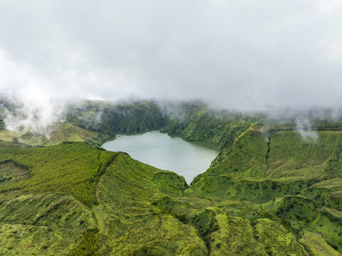 Aerial view of Miradouro Caldeira Rasa e Funda, a volcanic crater lake surrounded by lush green hills and low-hanging clouds in Mosteiro, Acores, Portugal.