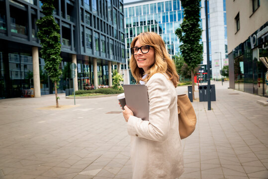 A blonde-haired woman walking through the business district with a laptop and a cup of coffee in her hands