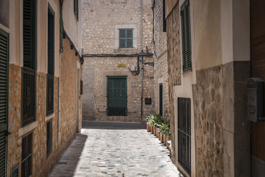 View of a narrow stone-paved street lined with historic stone buildings, green shutters, and potted plants in Soller, Balearic Islands, Spain.