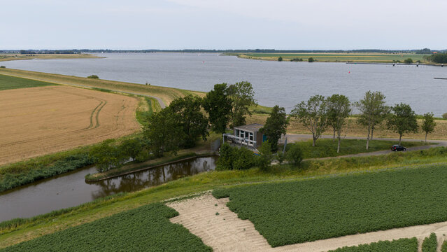 Aerial view of a small pumping station beside a canal, surrounded by agricultural fields and trees, with the Oosterschelde waterway in the background in Wilhelminadorp, Zeeland, Netherlands.