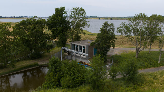 Aerial view of a small water management building situated next to a canal and a large body of water, surrounded by lush green trees and fields in Wilhelminadorp, Zeeland, Netherlands.