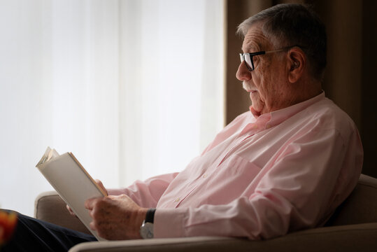 Close-up of a gray haired senior man sitting in an armchair and reading book in domestic room