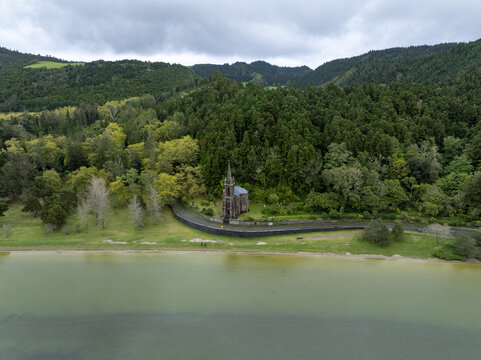 Aerial view of Ermida de Nossa Senhora das Vitorias situated on the shore of Lagoa das Furnas, surrounded by lush green forest and hills in Furnas, Acores, Portugal.