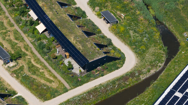 Aerial view of modern earth-sheltered houses with green roofs and integrated solar panels surrounded by lush vegetation and a small canal in Almere, Flevoland, Netherlands.