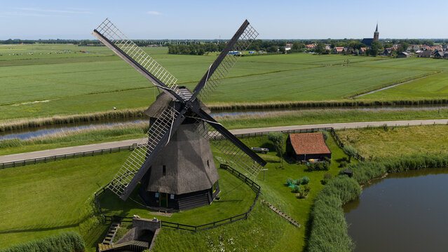 Aerial view of Bovenmolen G, a historic windmill with a thatched roof, set in a lush green landscape with canals and a small road in Schermerhorn, Noord-Holland, Netherlands.