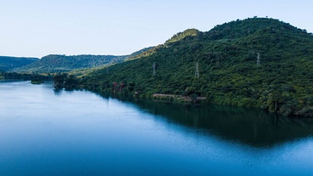 Aerial view of a wide river flowing past lush green hills with power transmission towers in Ghana.