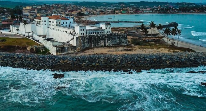 Aerial view of Elmina Castle, a historic white fortress situated on the coast with crashing waves, palm trees, and the town in the background, Ghana.