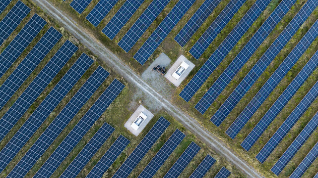 Aerial view of a large solar farm with rows of photovoltaic panels arranged in a grid pattern, separated by gravel paths and small electrical substations in Poland.