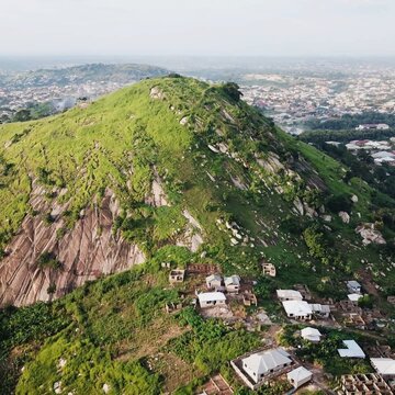 Aerial view of a lush green rocky hill with scattered houses at its base, surrounded by a sprawling urban landscape in Ghana.