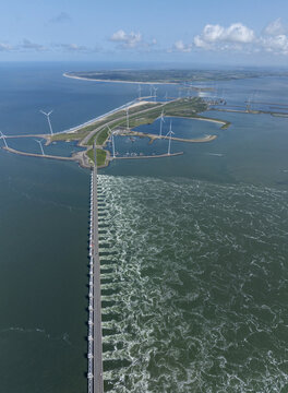 Aerial view of the Eastern Scheldt Storm Surge Barrier with turbulent water flowing through the gates, wind turbines, and coastal landscape in Vrouwenpolder, Zeeland, Netherlands.