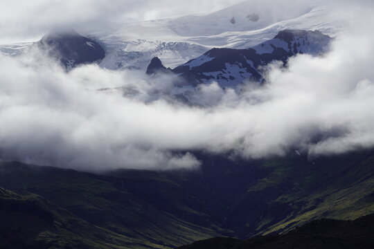 View of Skaftafell mountain range with snow-capped peaks partially obscured by low-hanging clouds and lush green slopes in Skaftafell, Sveitarfelagid Hornaf, Iceland.