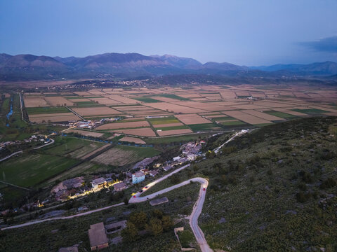Aerial view of the agricultural landscape with a winding road illuminated by car lights at dusk, set against a backdrop of mountains in Saranda, Vlore County, Albania.