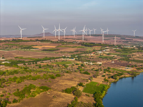 Aerial view of Phuoc Dinh Wind Farm with numerous wind turbines set against a landscape of dry fields, agricultural plots, and a reservoir in Phuoc Dinh, Ninh Thuan, Vietnam.
