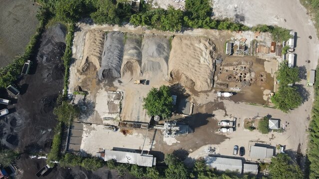 Aerial view of an industrial construction site featuring large piles of sand and gravel, concrete mixers, and trucks parked on a dirt lot in Cilegon, Banten, Indonesia.