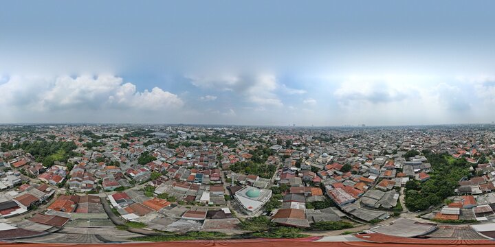 Aerial view of the urban landscape of Depok featuring dense residential rooftops, lush green trees, and a mosque dome under a clear blue sky in Depok, West Java, Indonesia.