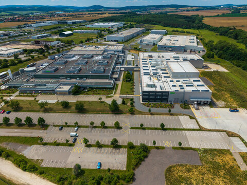 Aerial view of a modern industrial park with large warehouse buildings, flat roofs, parking lots, and surrounding green fields in Zebrak, Central Bohemian Region, Czechia.