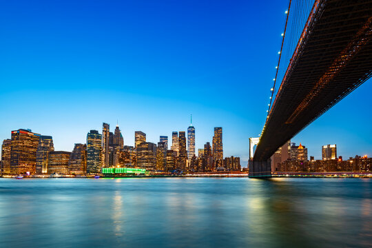 Spectacular view of downtown Manhattan with the iconic Brooklyn Bridge stretching across the water as city lights reflect on the river.