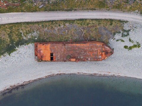 Aerial view of a rusted shipwreck resting on a rocky beach near the shoreline in Unalaska, Alaska, United States.